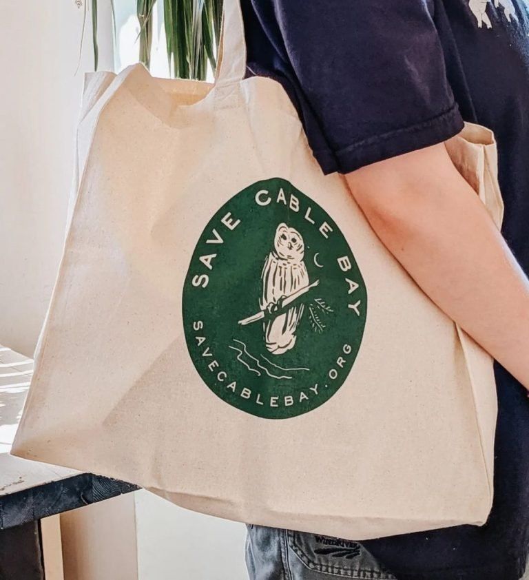 A person holding a canvas tote bag printed with the Save Cable Bay otter logo