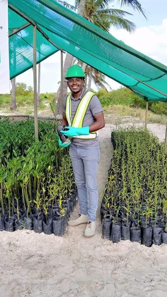 A smiling man in a hard hat and hi-vis vest among nursery seedlings, holding a phone