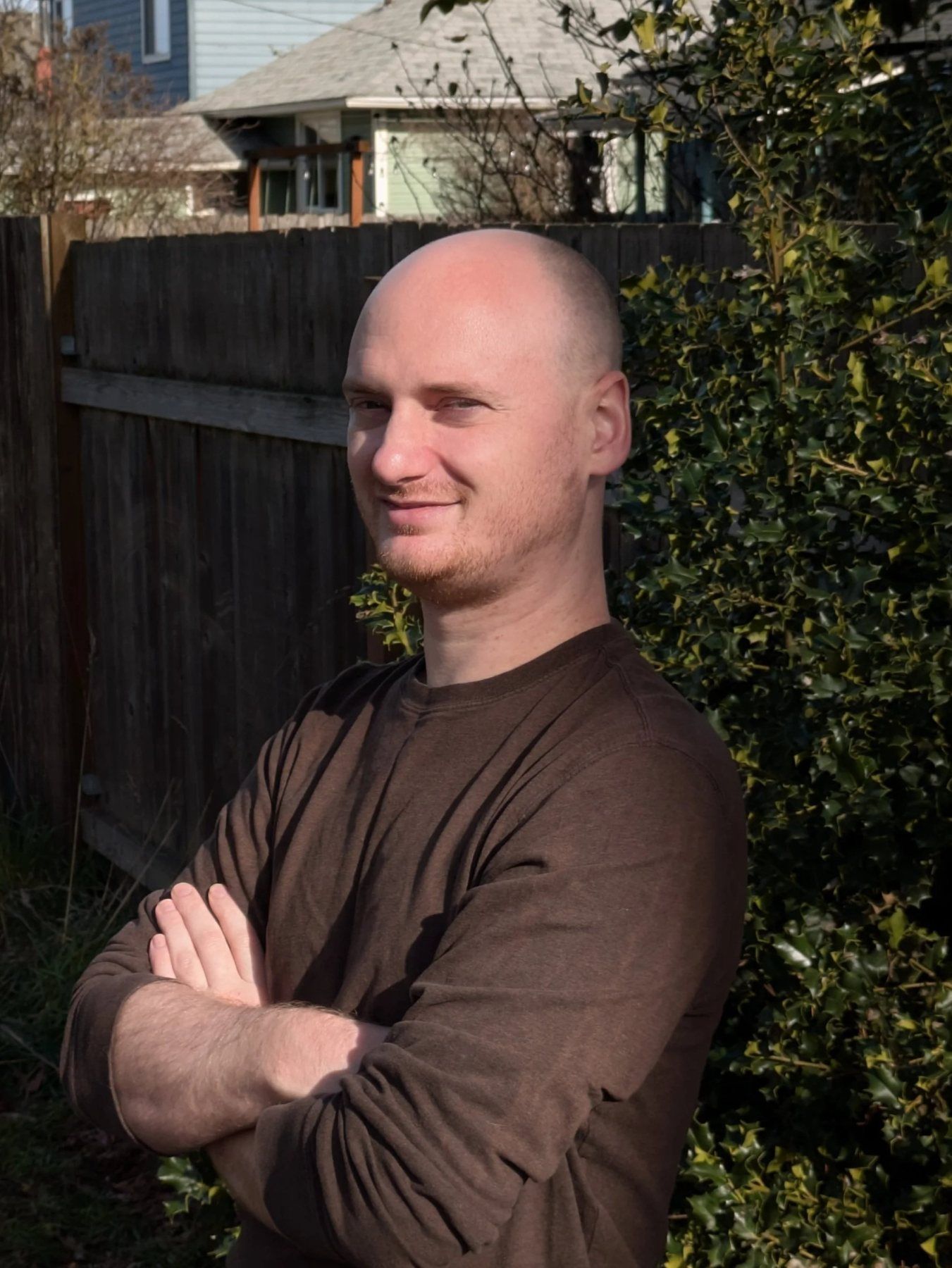 A man with a shaved head and short beard standing outdoors with arms crossed