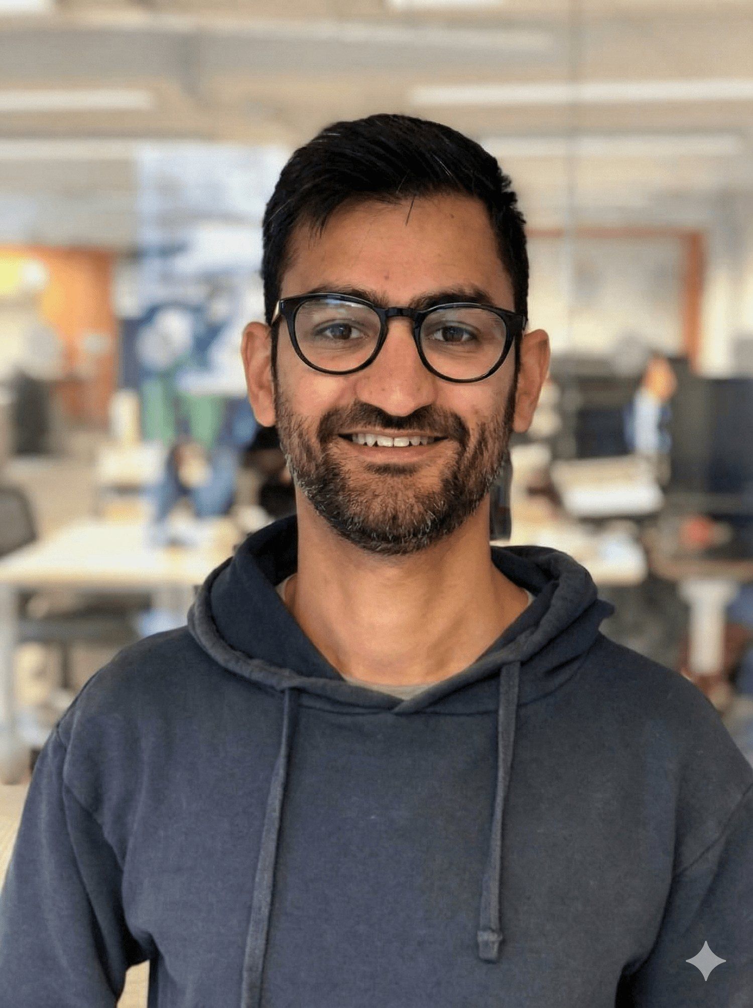 Samir Gandhi smiling in an office, wearing glasses and a dark hoodie